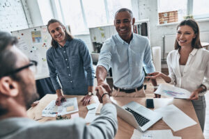 Top view of young modern men in smart casual wear shaking hands and smiling while standing with their colleagues in the board room.